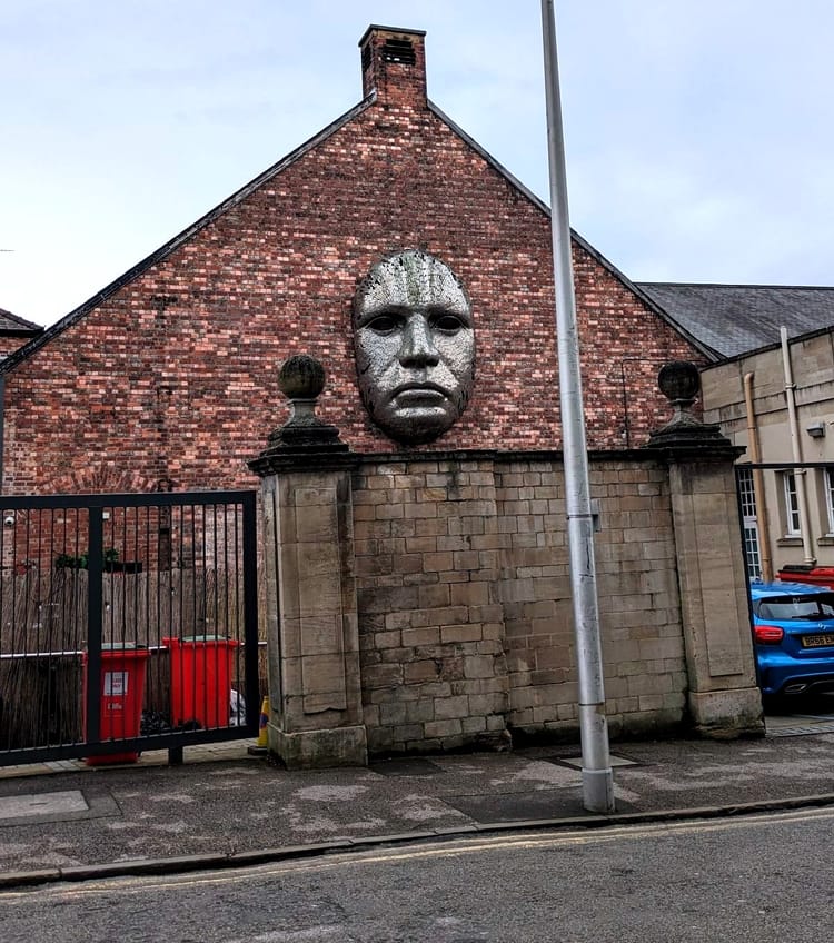 A large metallic face protrudes from the exterior of a red brick wall